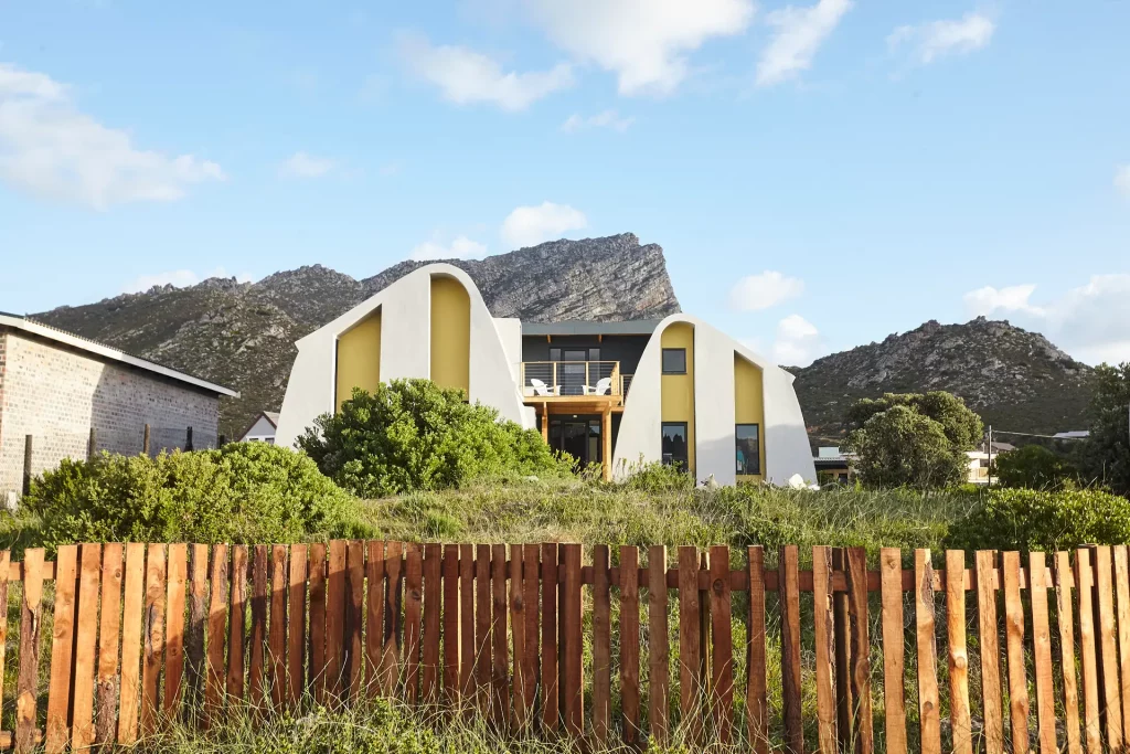 House Mahika, a sustainable, modern villa with a curved roof, positioned in a landscape of green grass and a blue sky, with a wooden fence in the foreground and cliffs in the background.